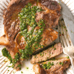 A piece of cooked Denver steak on a white plate with chimichurri sauce on it.