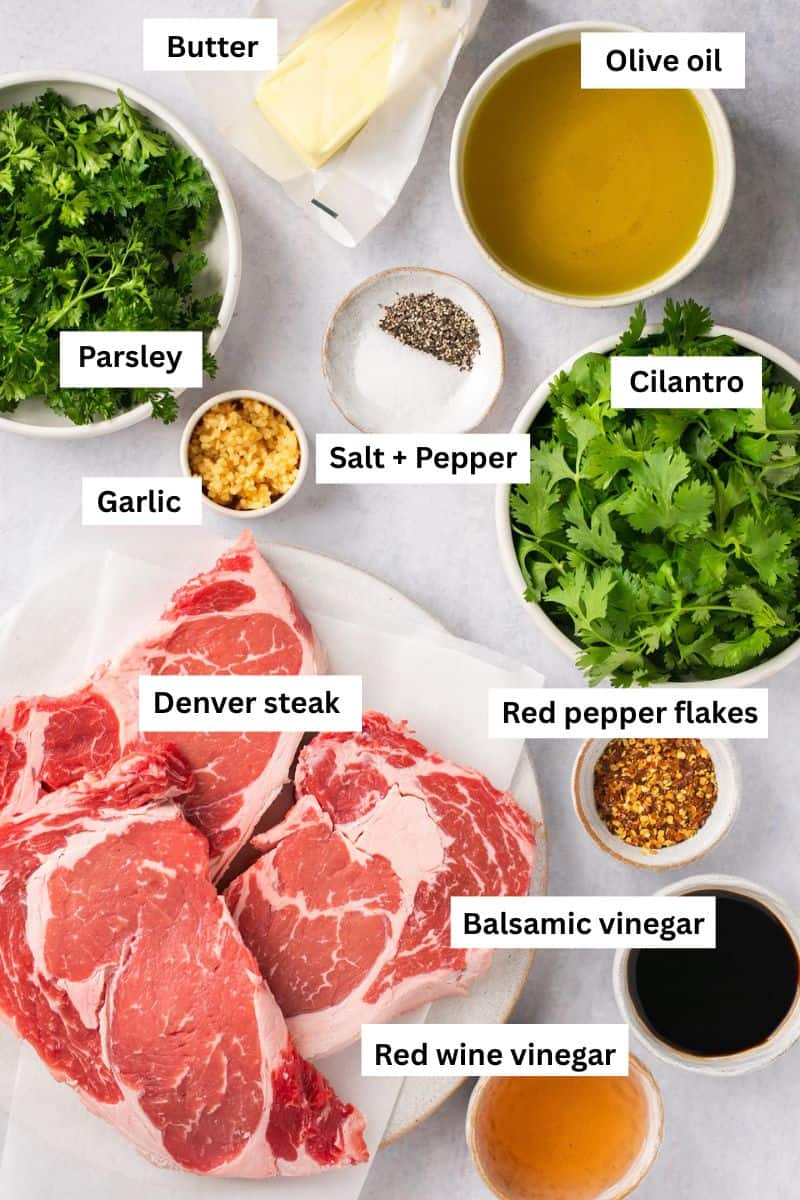 A plate with uncooked steak on it surrounded by fresh herbs, a stick of butter, salt and pepper and crushed garlic.