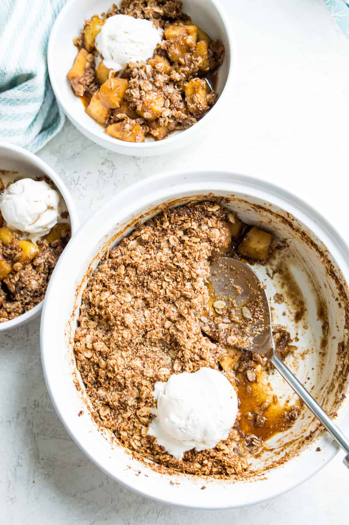 A casserole dish filled with a health apple crumble with ice cream on top and a serving spoon in it.