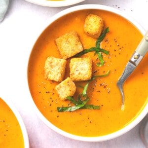 A bowl of butternut squash and red pepper soup garnished with croutons and fresh herbs.