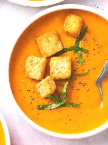 A bowl of butternut squash and red pepper soup garnished with croutons and fresh herbs.