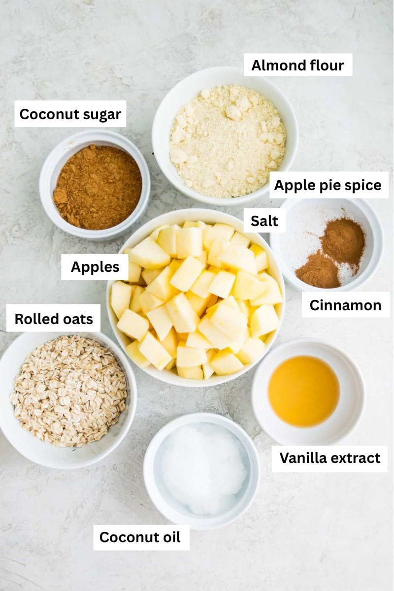 The ingredients needed to make apple crumble separated into bowls including peeled apples, sugar, ground cinnamon, rolled oats and flour.