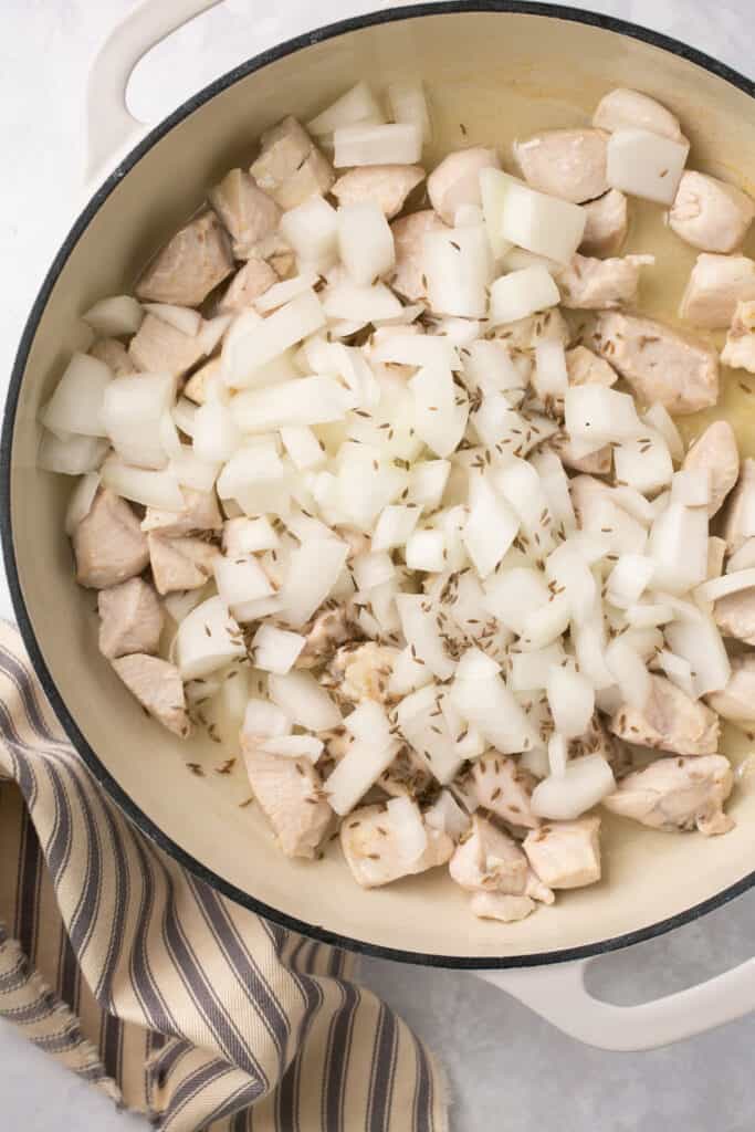 A pan with cooked chicken chunks, raw chopped onion and cumin seeds in it on the stovetop.