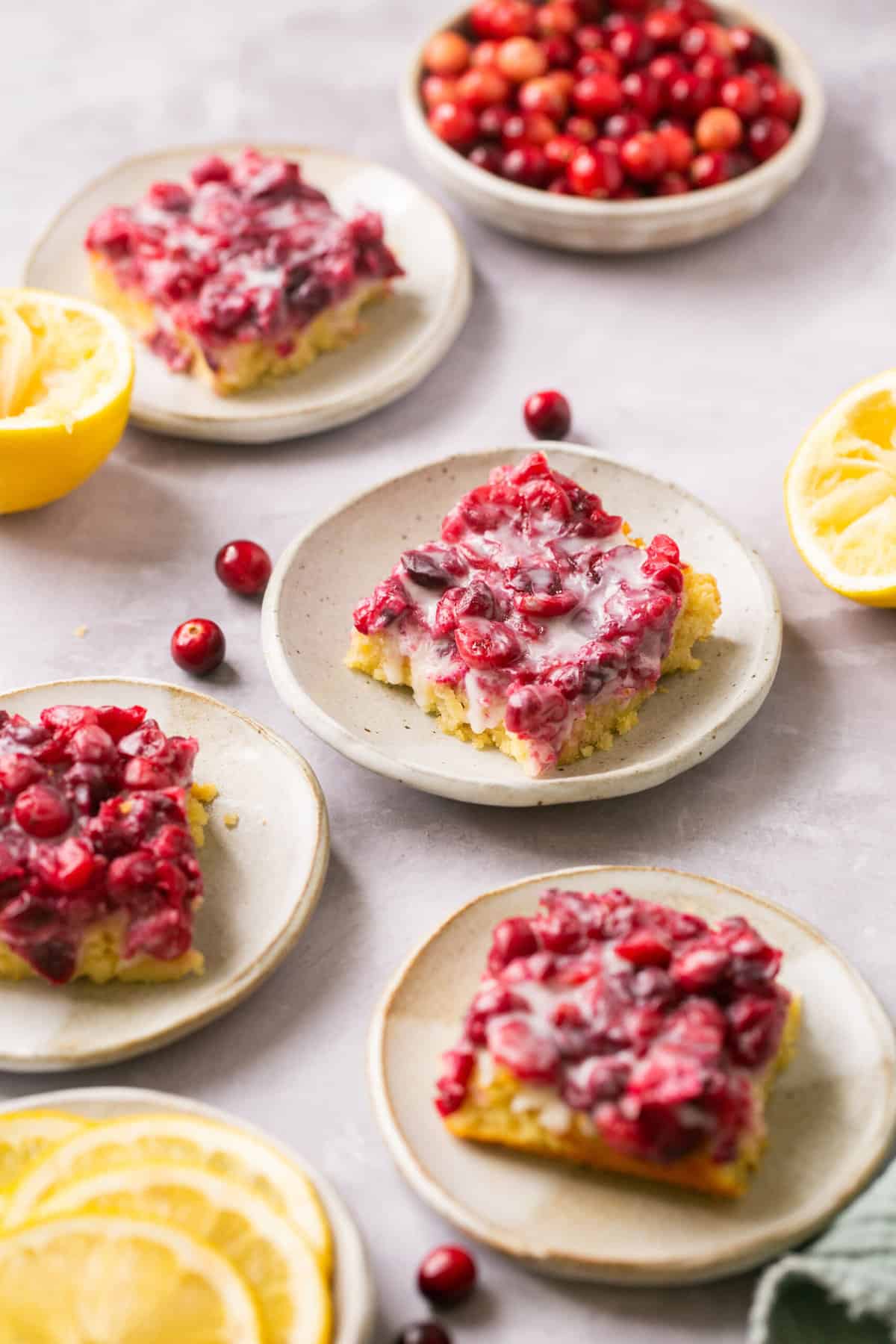 Four serving plates of paleo cranberry lemon bars alongside bowls of cranberries and orange slices.