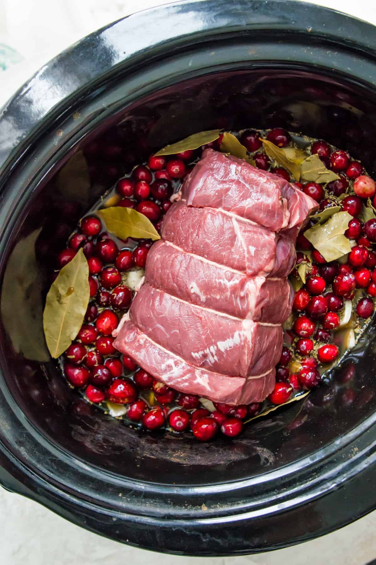 A beef roast in a slow cooker bowl surrounded by cranberries and bay leaves. 