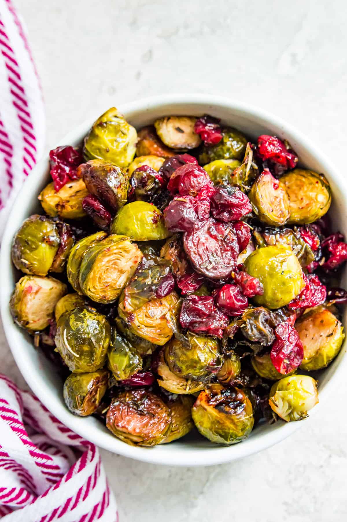 A bowl or roasted Brussels sprouts with cranberries and herbs and a tea towel beside it.