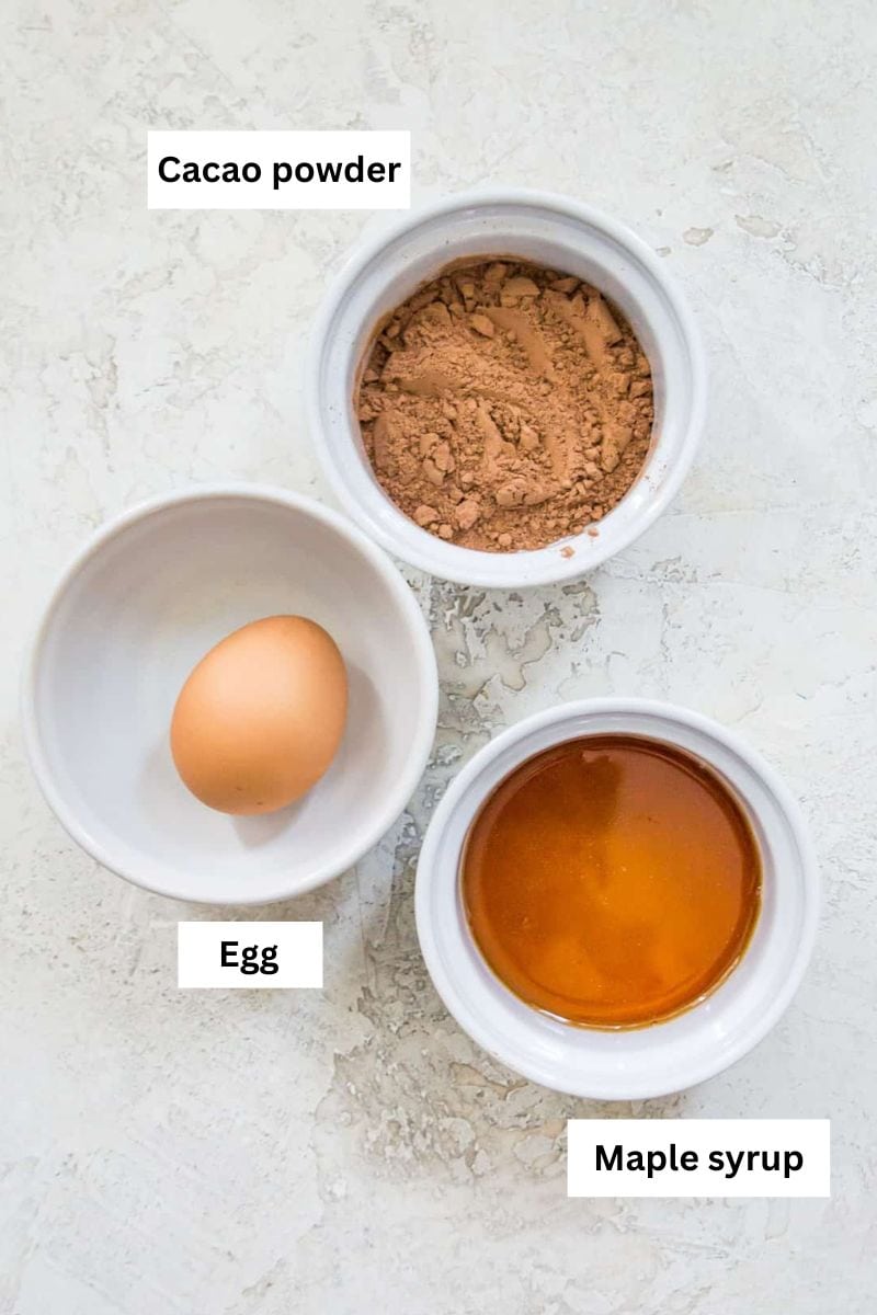 The ingredients needed to make a mug cake separated into bowls including cacao powder, an egg and maple syrup.