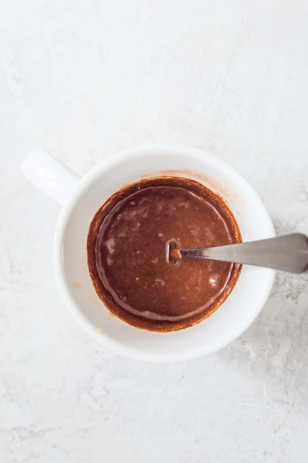 A mug with chocolate cake batter in it and a spoon in the batter.