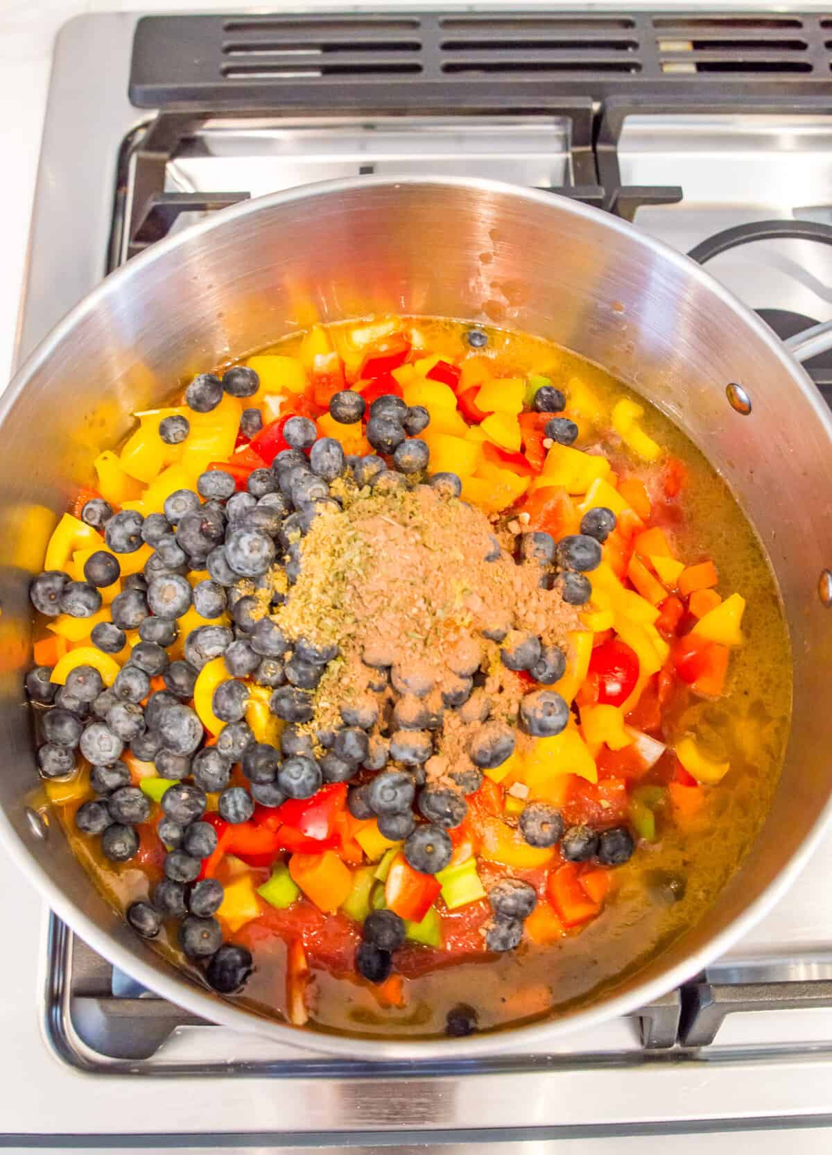 A pot filled with chopped vegetables, cooked ground beef, blueberries, spices and broth.