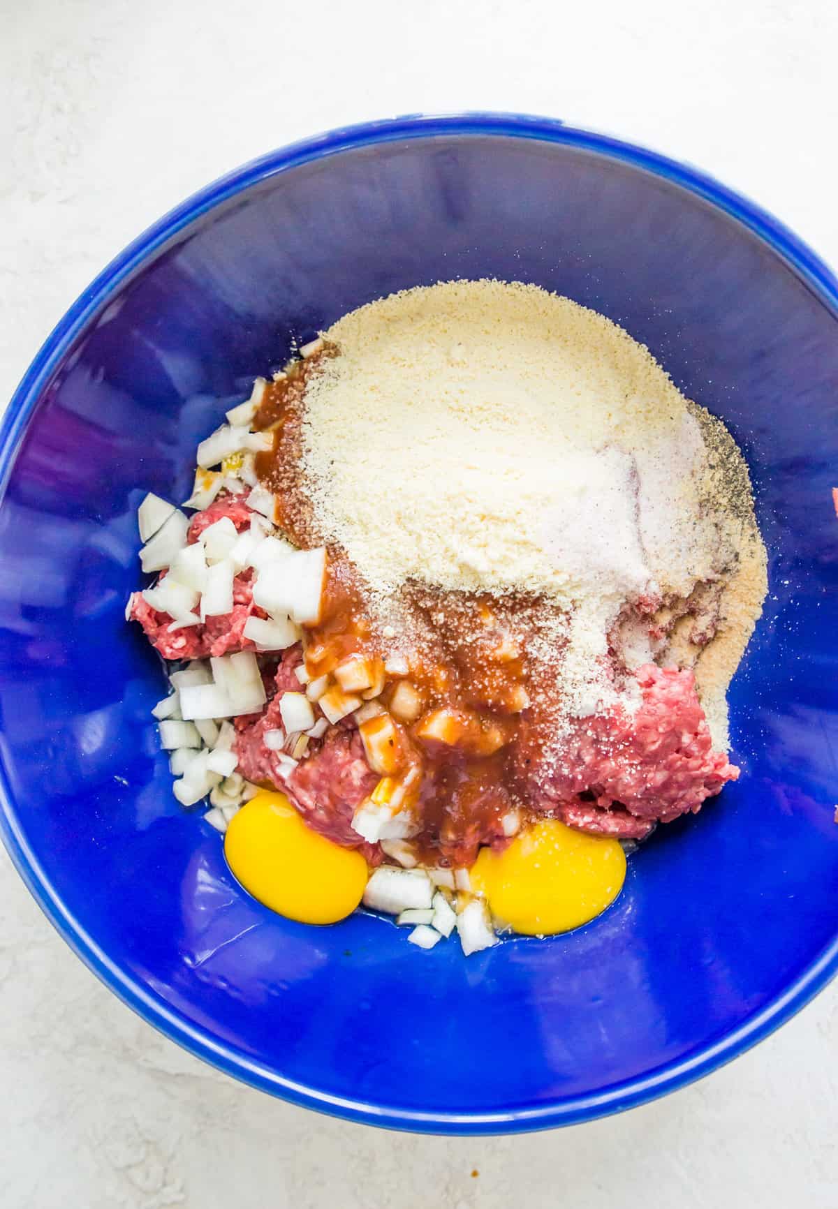 A large blue bowl filled with ground beef, almond flour, chopped onions, barbecue sauce and eggs.