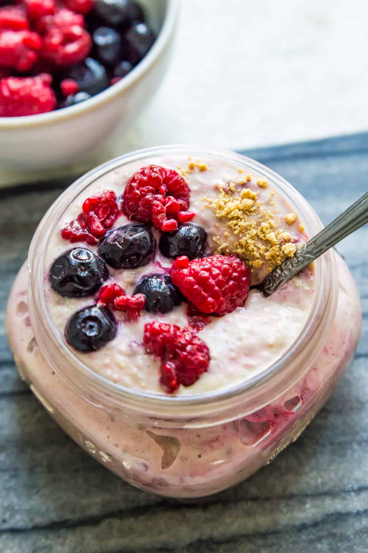 A jar of berry cheesecake overnight oats topped with fresh raspberries and blueberries with a spoon in it.