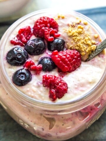 A jar of berry cheesecake overnight oats topped with fresh berries with a spoon in it.