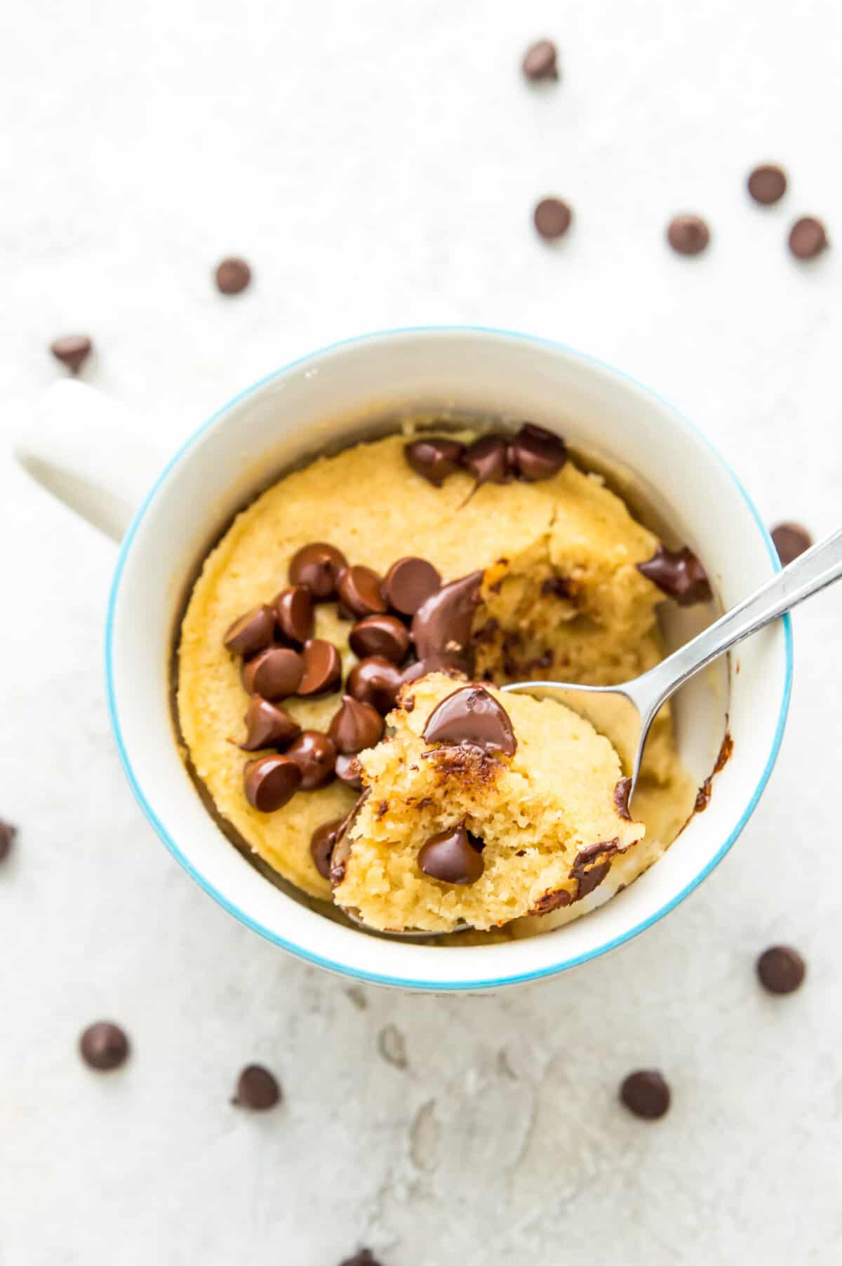 A mug of vanilla protein mug cake topped with chocolate chips with a spoon in it.