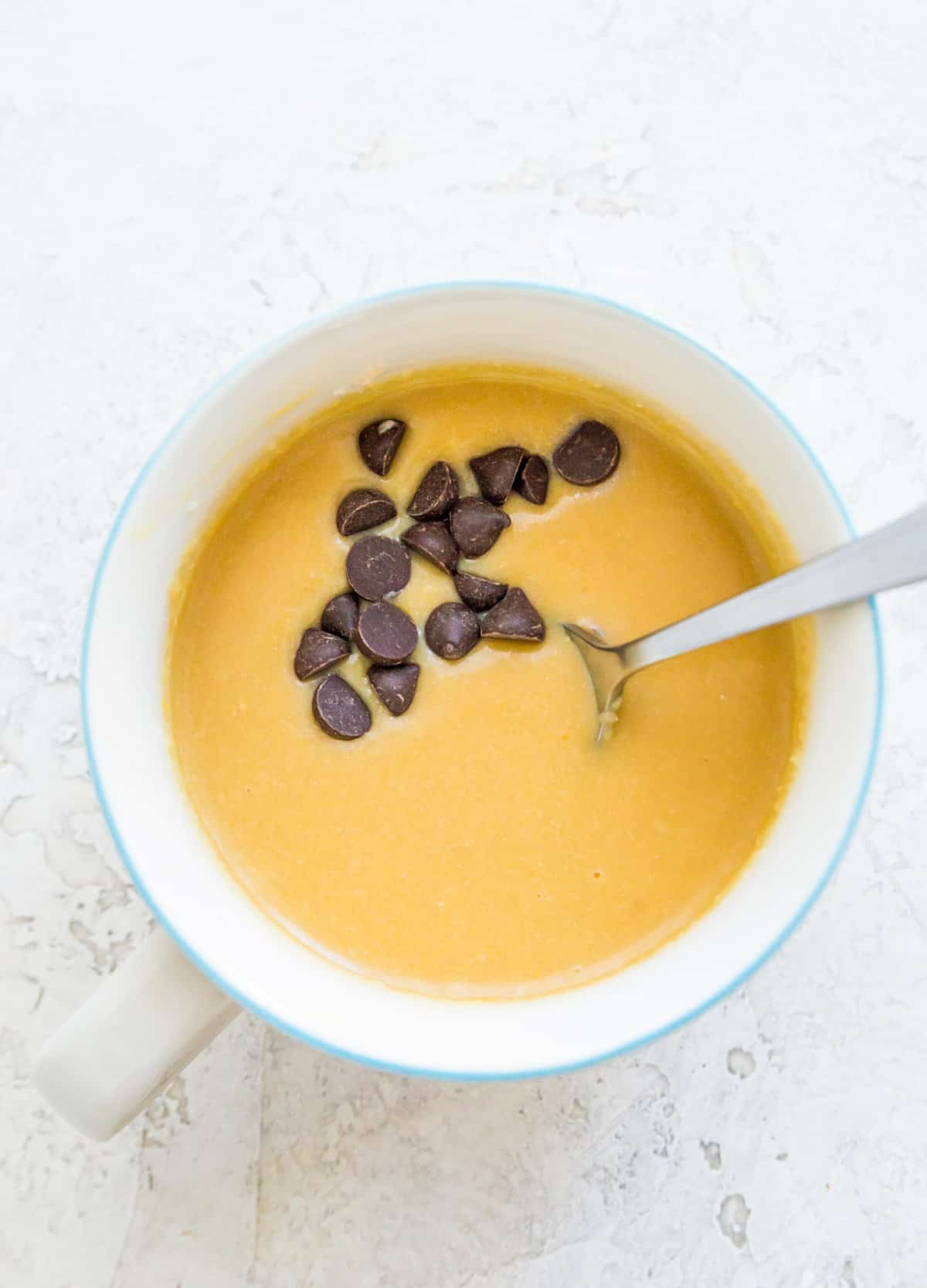 Vanilla protein mug cake batter in a mug with a spoon in it and topped with chocolate chips. 