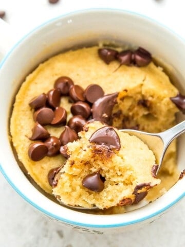 A mug of a vanilla protein mug cake with chocolate chips on top of it and a spoon in it.