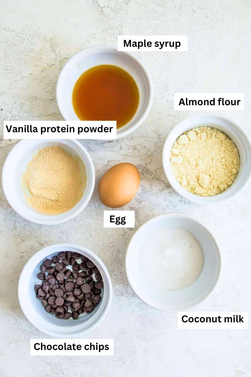 The ingredients for making a mug cake separated into bowls including an egg, maple syrup, almond flour, protein powder and coconut milk.