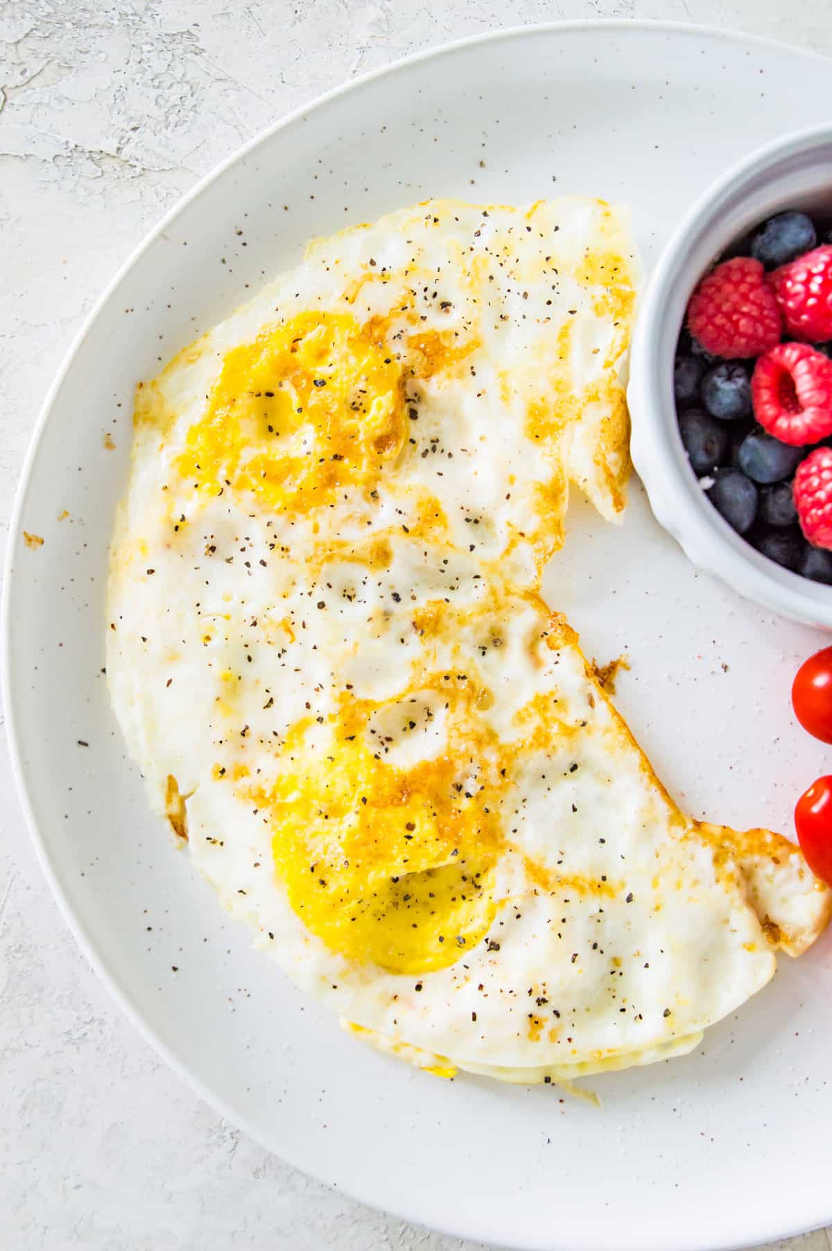 Two over hard eggs on a plate with a side bowl of fresh blueberries and raspberries. 