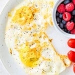 A white plate with over hard eggs on it and a side dish full of fresh raspberries and blueberries.