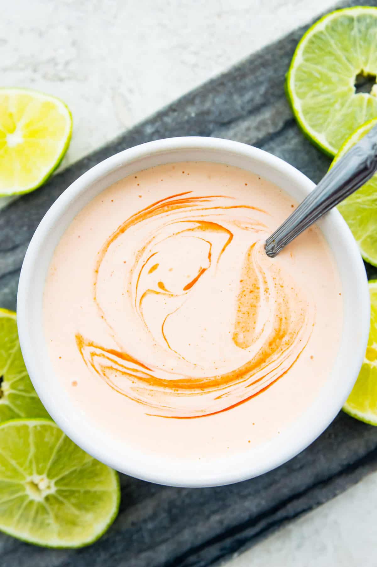 A bowl of sriracha aioli with a spoon in it surrounded by lime slices. 