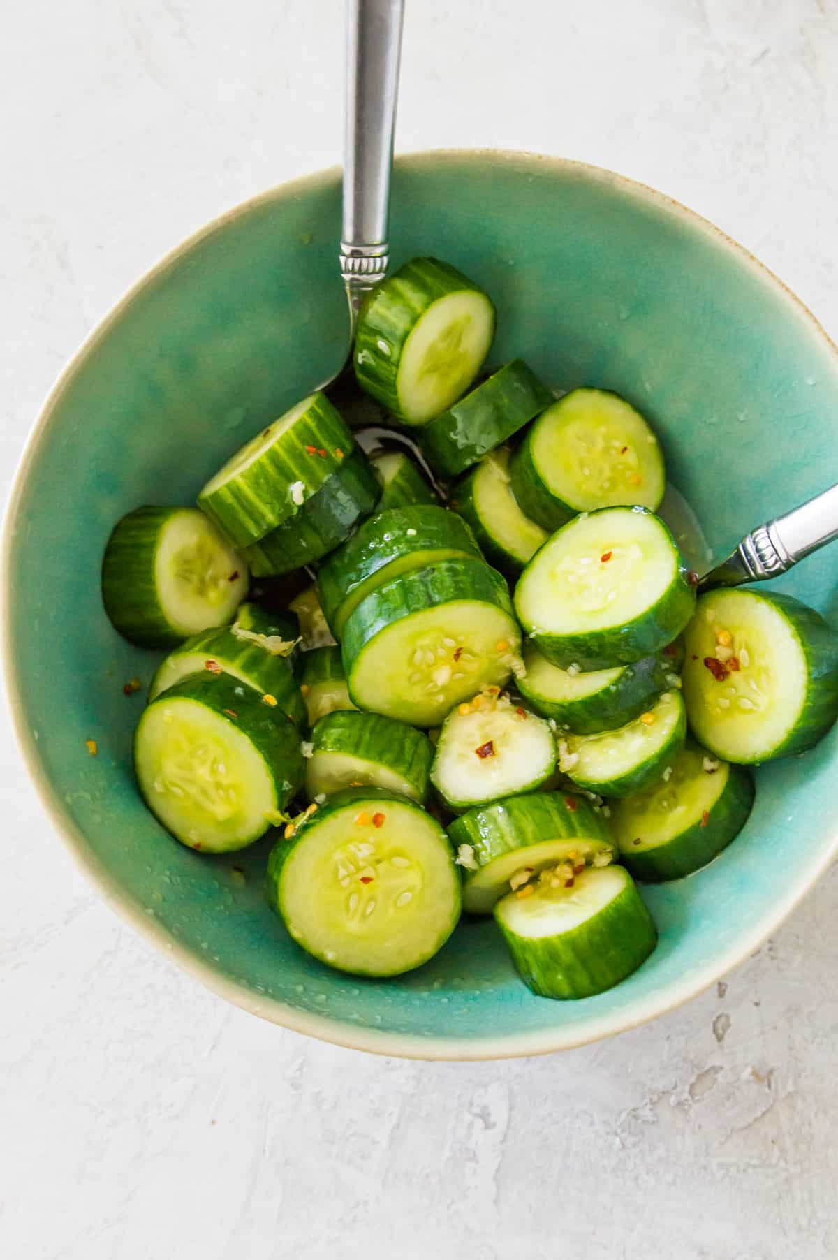 Sliced cucumbers in a bowl with a dressing on them and spoons in the bowl.