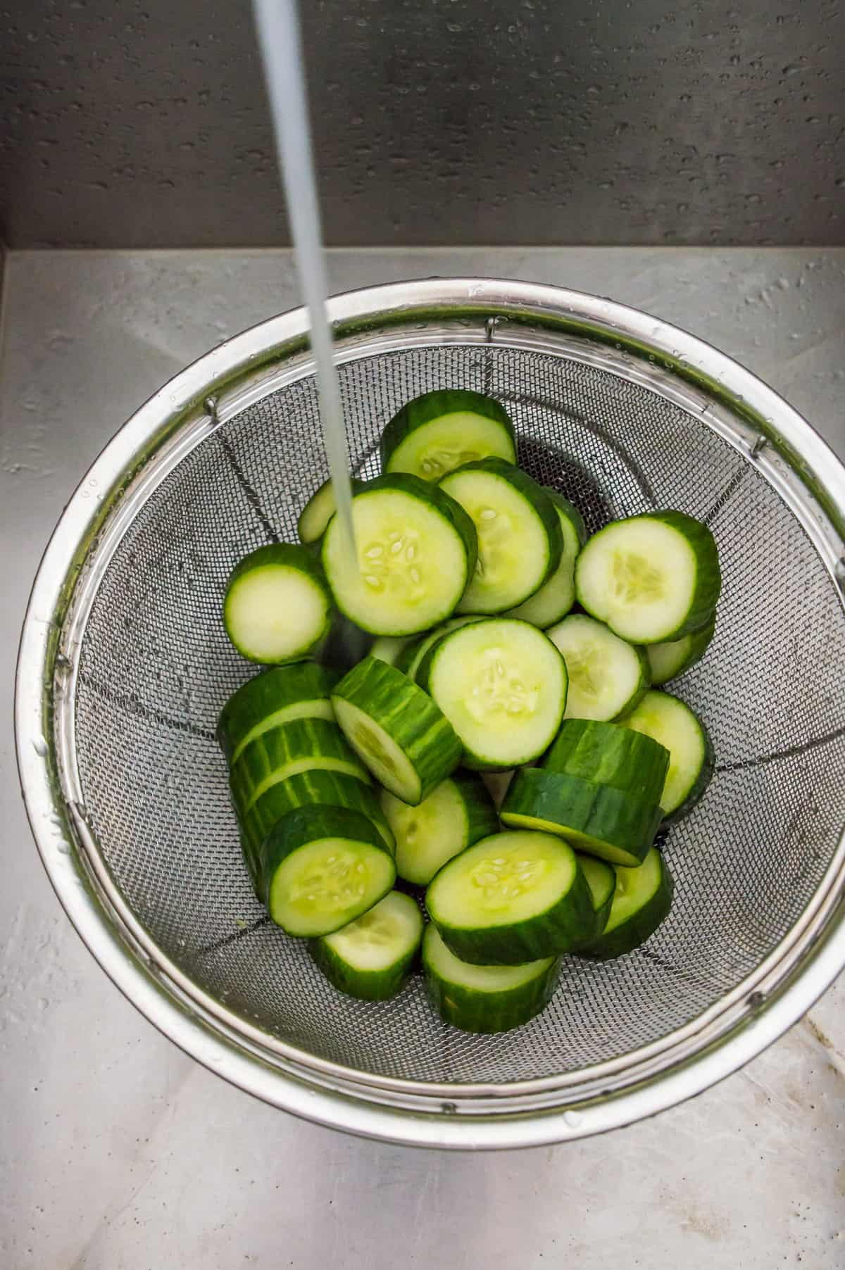 Sliced cucumbers in a colander with water being poured on them.