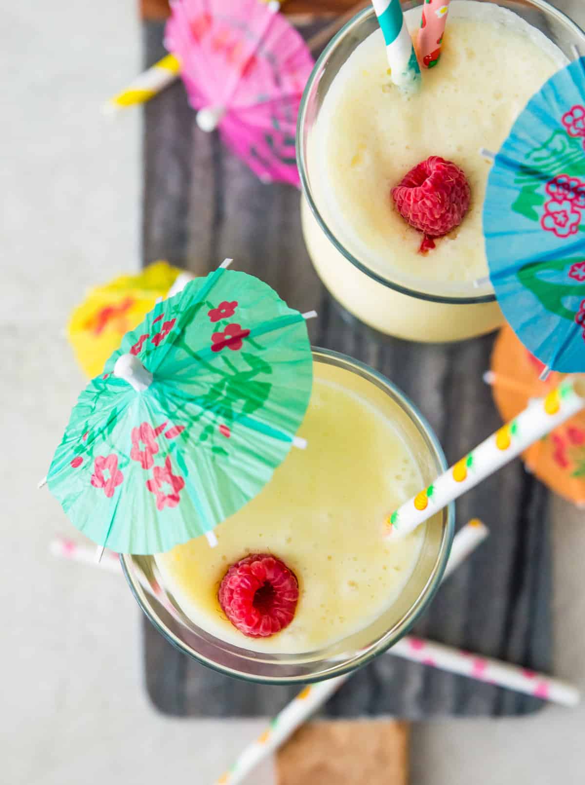 Two glasses of virgin piña coladas topped with fresh raspberries, drink umbrellas and straws. 