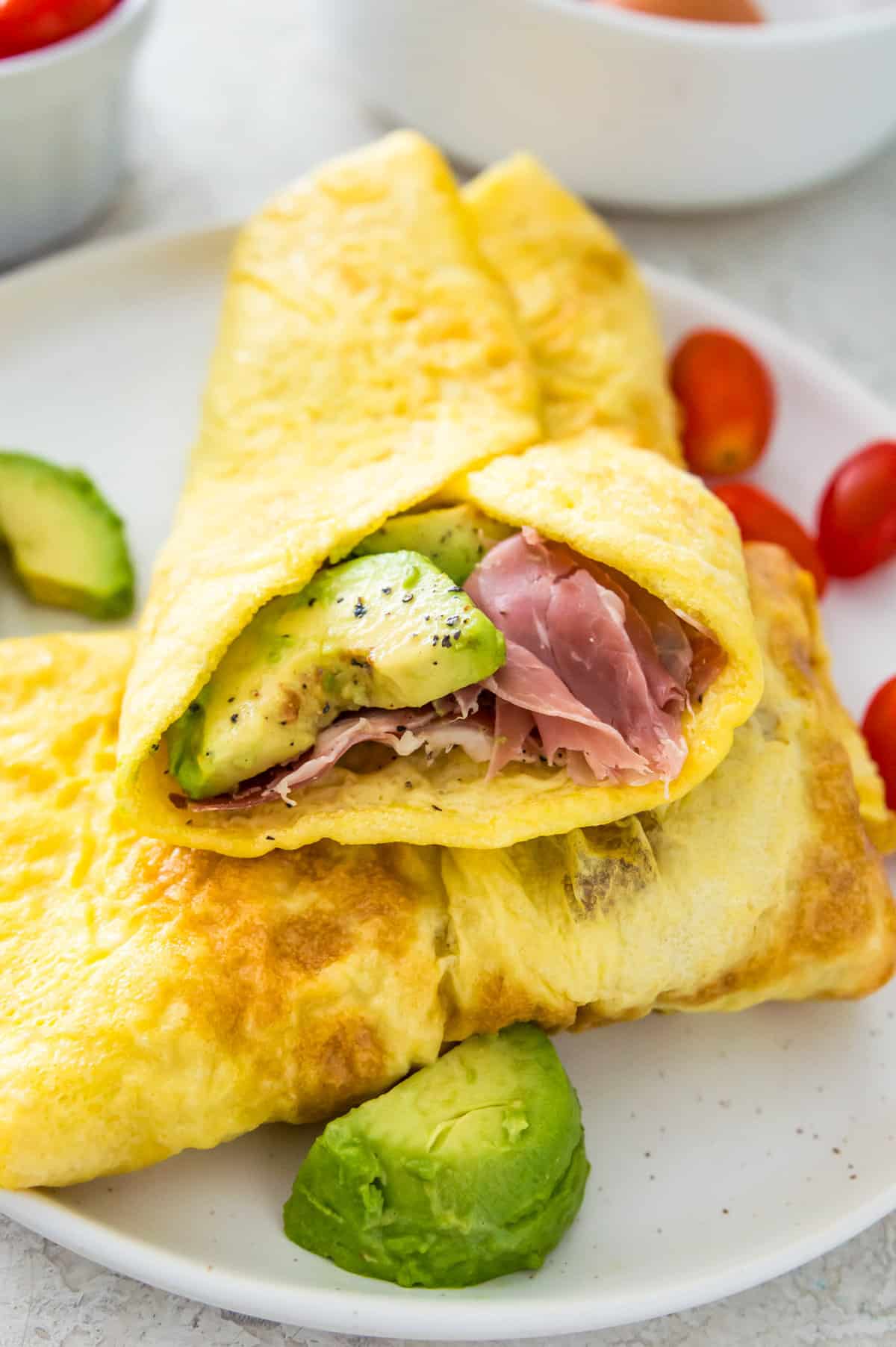 An egg wrap filled with sliced avocado and ham on a plate with baby tomatoes.