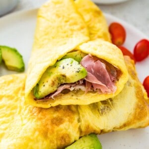 An egg wrap with sliced avocado and ham in it on a plate with baby tomatoes beside it.