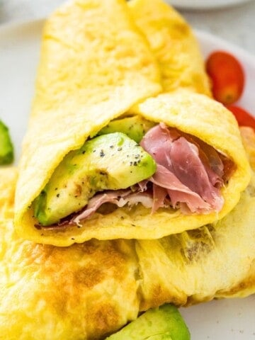 An egg wrap with sliced avocado and ham in it on a plate with baby tomatoes beside it.