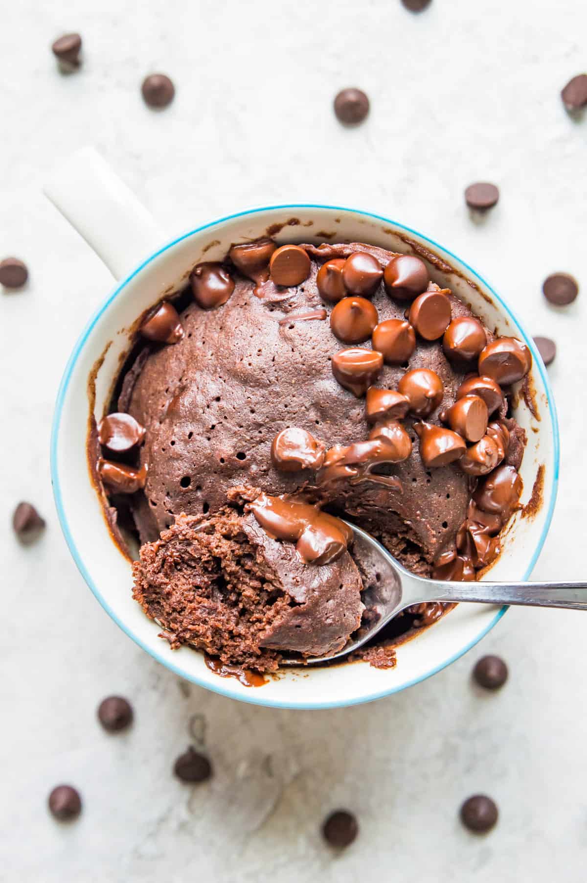 A chocolate mug cake with chocolate chips on top of it and a spoon with a piece of cake resting on it.