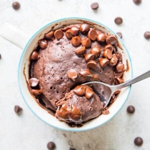 A mug full of a chocolate gluten free mug cake topped with chocolate chips and with a spoon in it.