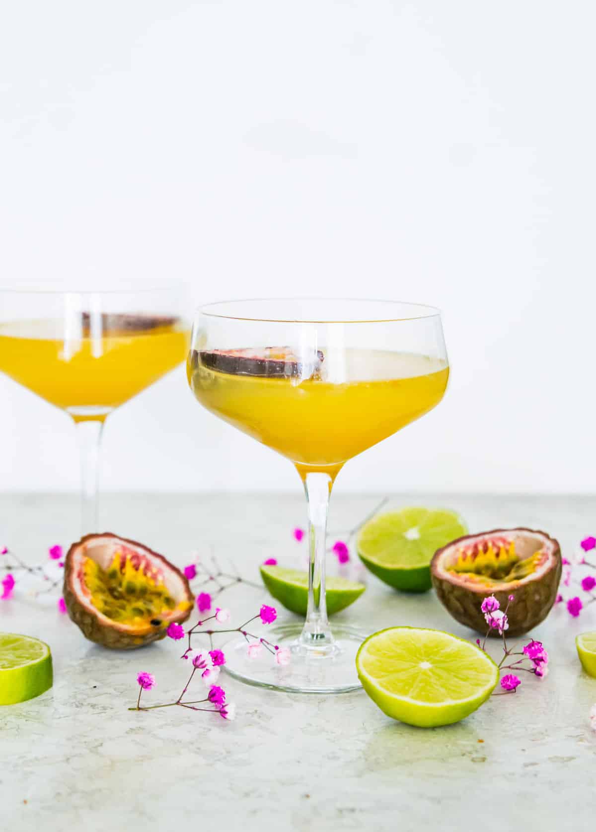 Two pornstar martini mocktails in martini glasses surrounded by pink flowers and lime slices.