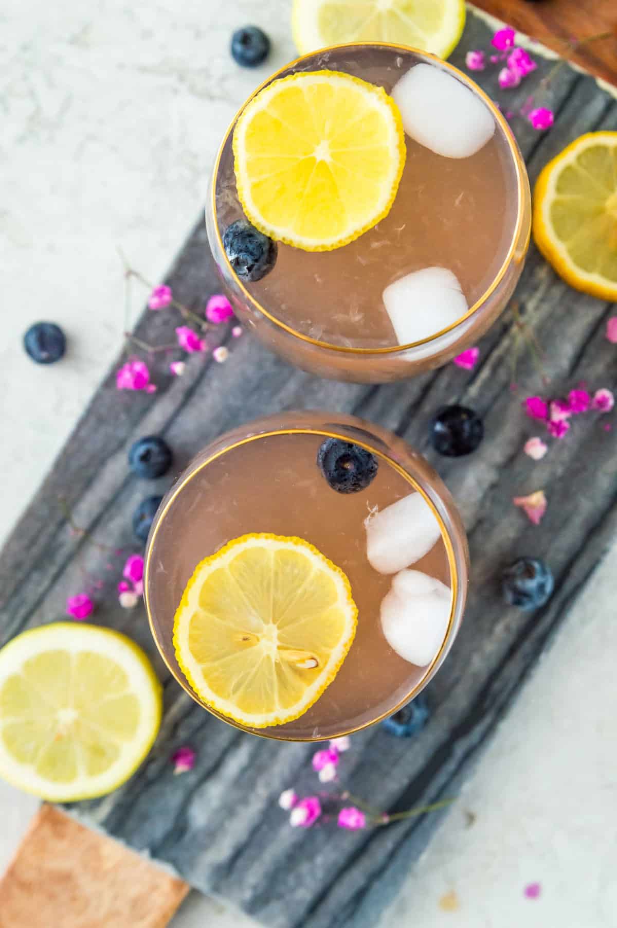 Two glasses of huckleberry lemonade topped with lemon slices, fresh blueberries and ice. 