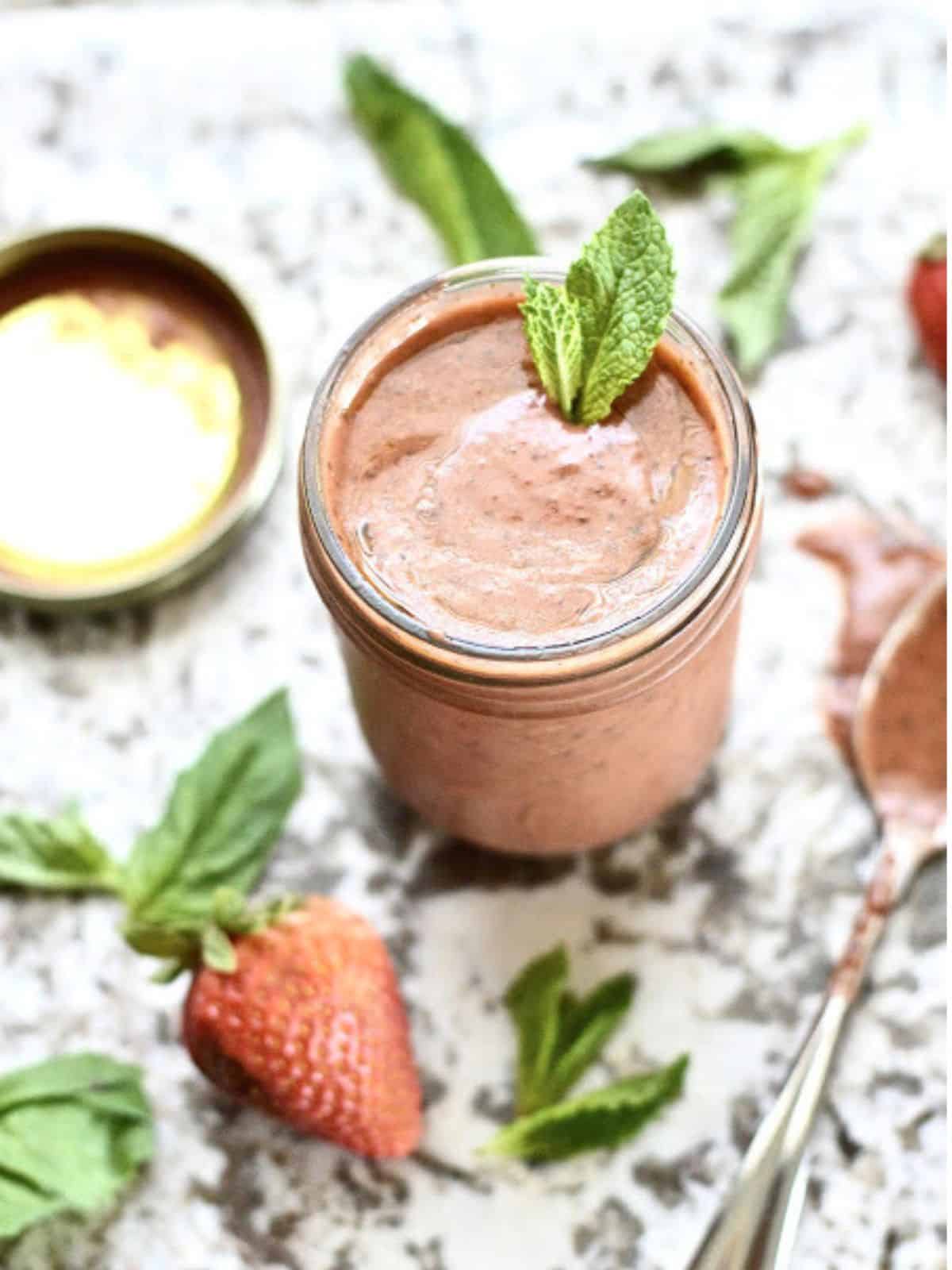 A jar of strawberry vinaigrette topped with fresh mint leaves with a spoon beside it and strawberries around it.