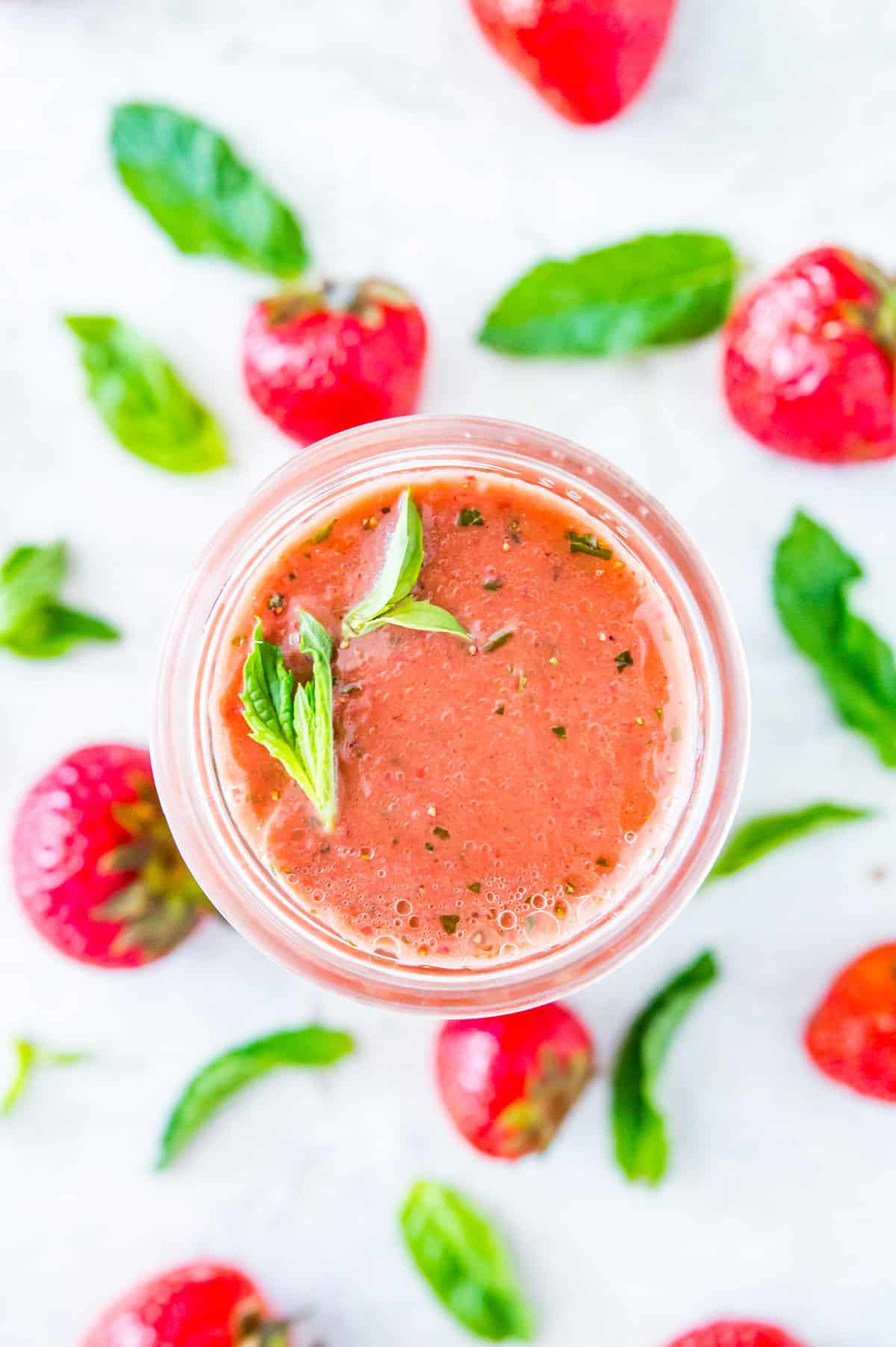 A jar of strawberry basil salad dressing topped with fresh mint leaves surrounded by strawberries and basil leaves.