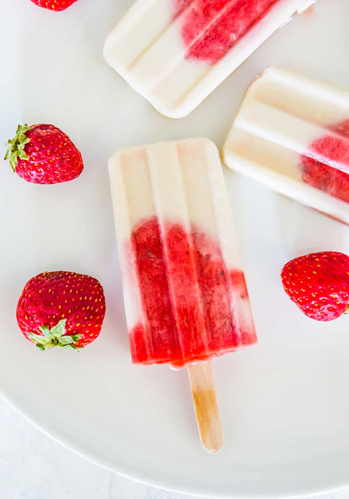 Three strawberry and vanilla popsicles on a white plate surrounded by fresh strawberries.