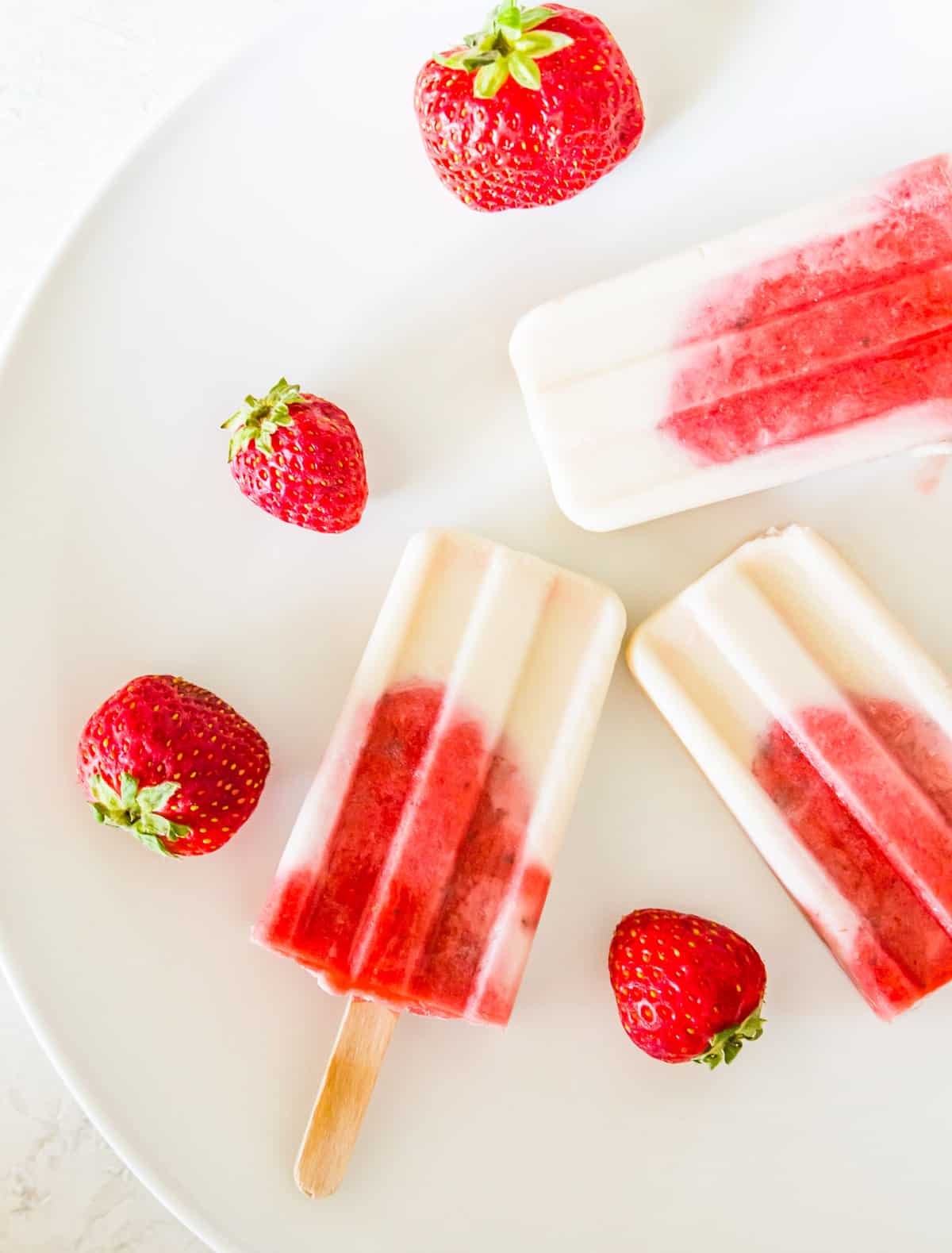 Three strawberry vanilla popsicles on plate with fresh strawberries around them.