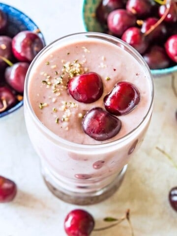A clear glass with a cherry smoothie in it, topped with fresh cherries and hemp hearts with fresh cherries around the glass.