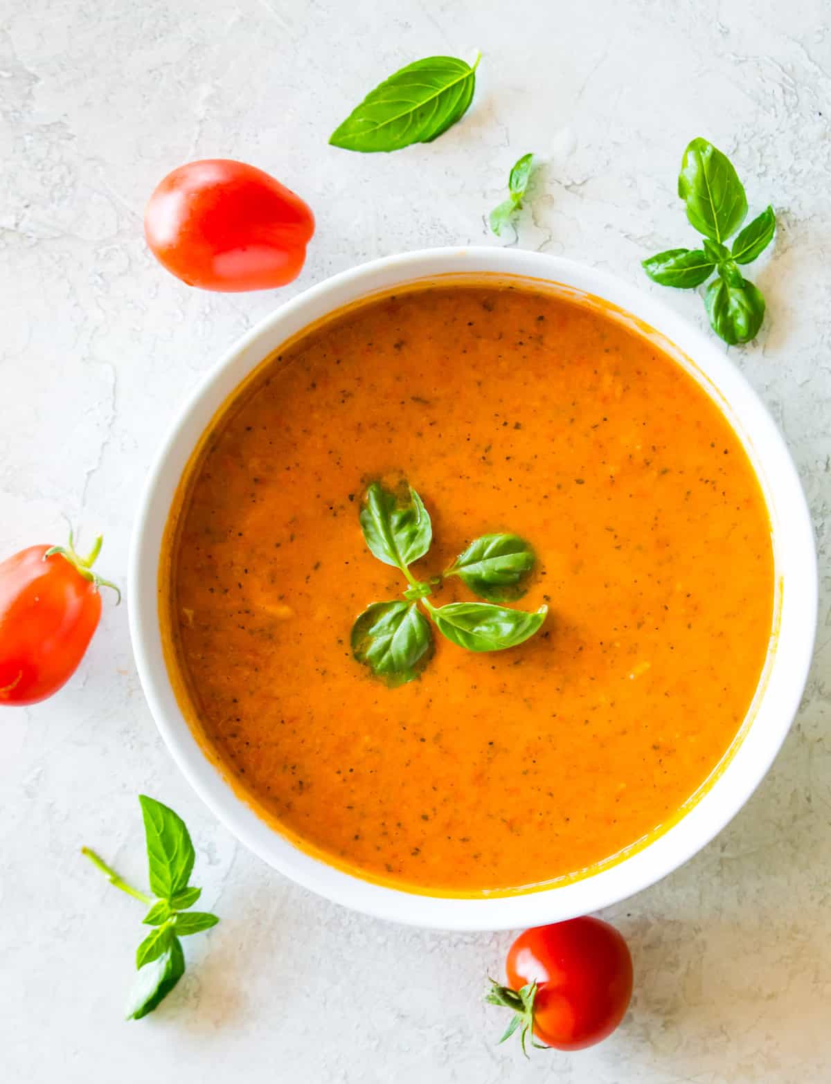 A bowl of tomato soup topped with fresh basil and surrounded by basil and tomatoes. 