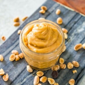 A small, clear jar filled with peanut butter mousse on a serving tray surrounded by peanuts.
