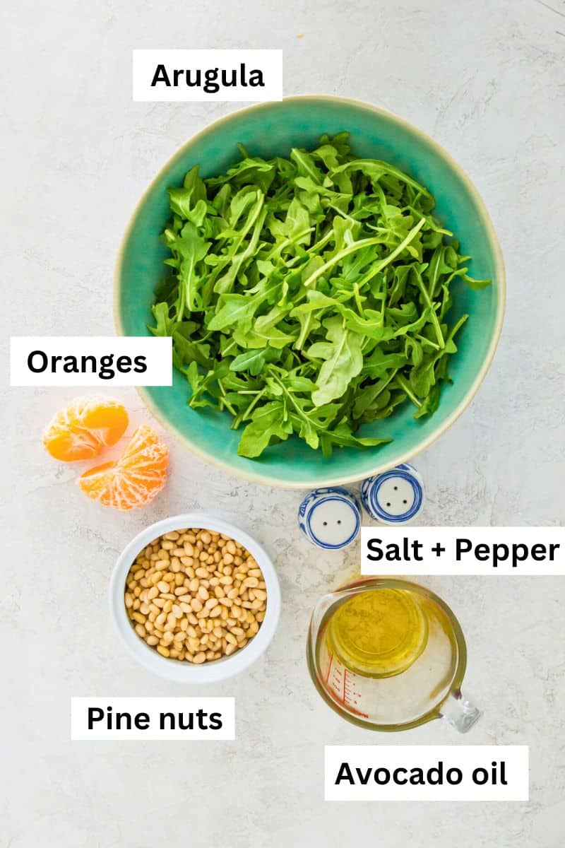 The ingredients needed to make arugula pesto separated into bowls including fresh arugula, mandarine oranges, pine nuts, avocado oil and salt and pepper.