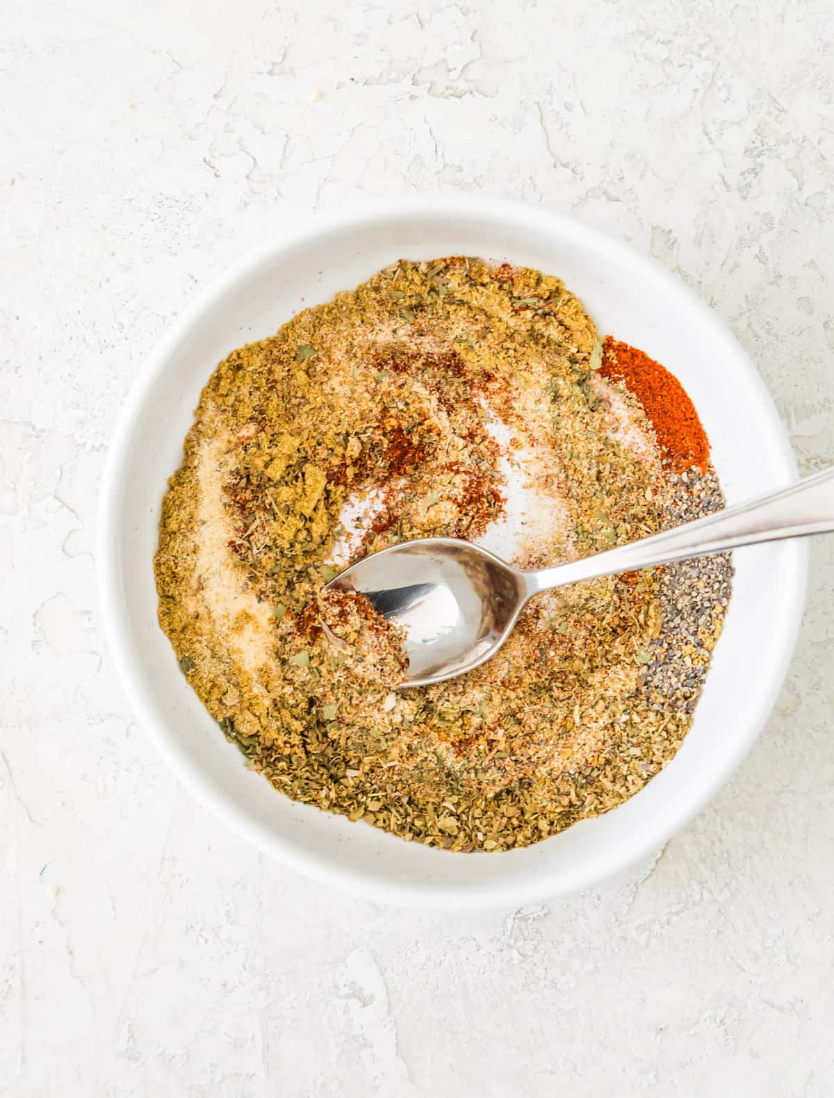 Different dried herbs and spices in a white bowl with a spoon in it.