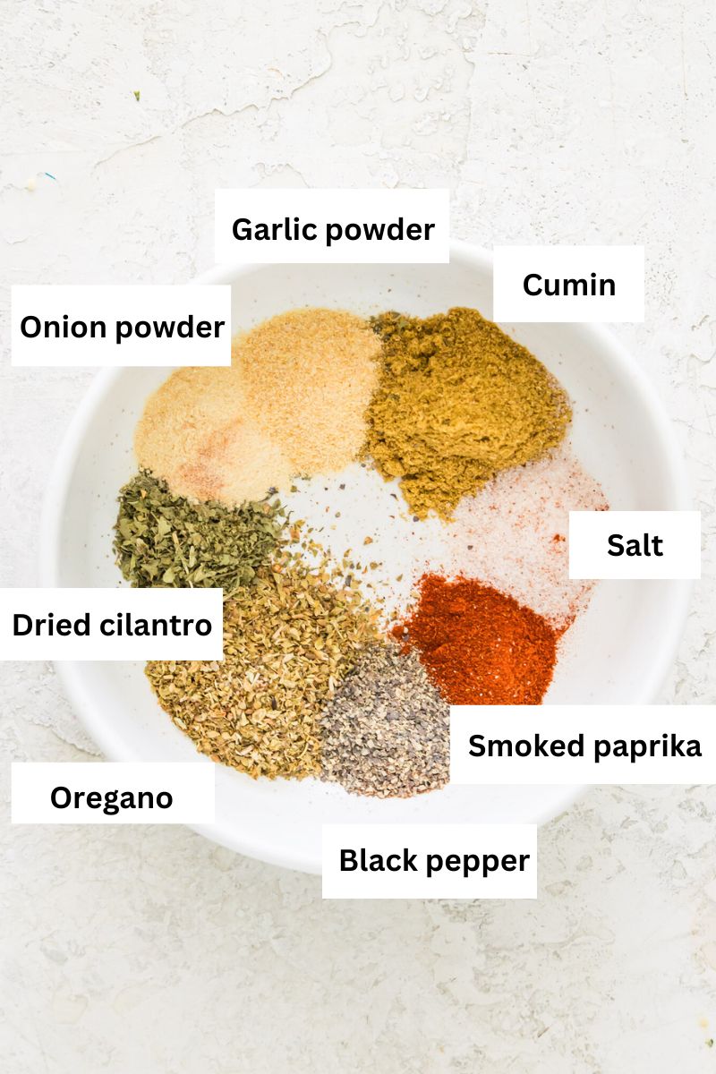 A white bowl filled with the spices needed to make a taco seasoning mix including cumin, paprika, onion powder, garlic powder, salt, pepper and oregano.
