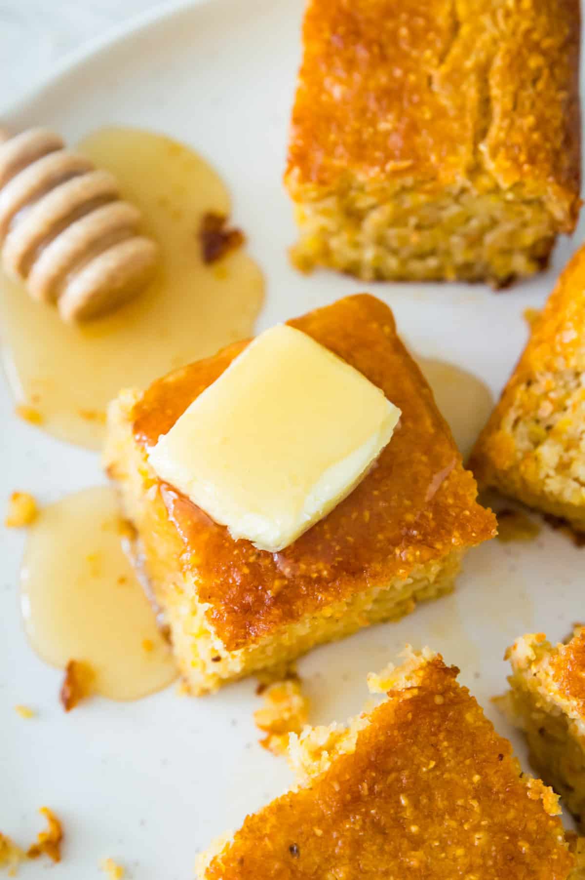 A white plate with pieces of gluten free cornbread on it, and one of them is topped with a square of butter and has honey drizzled on it.