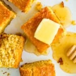 A white plate with pieces of cornbread on it, with one of them topped with a square of butter and drizzled in honey.