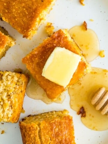 A plate with many pieces of cornbread on it, one of them with a slab of butter on top with a drizzle of honey on it.