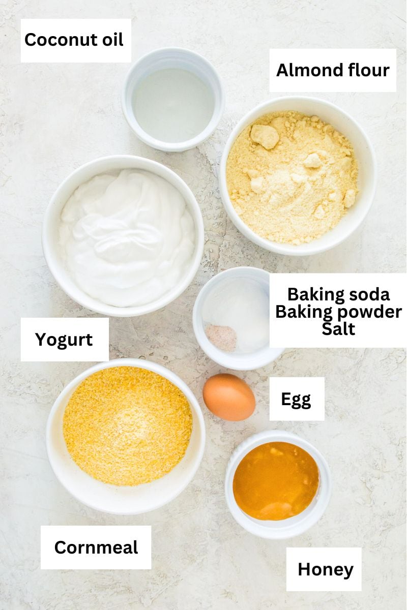 Small bowls filled with the ingredients for making cornbread including almond flour, cornmeal, yogurt, egg, baking soda, baking powder, salt, honey and coconut oil.