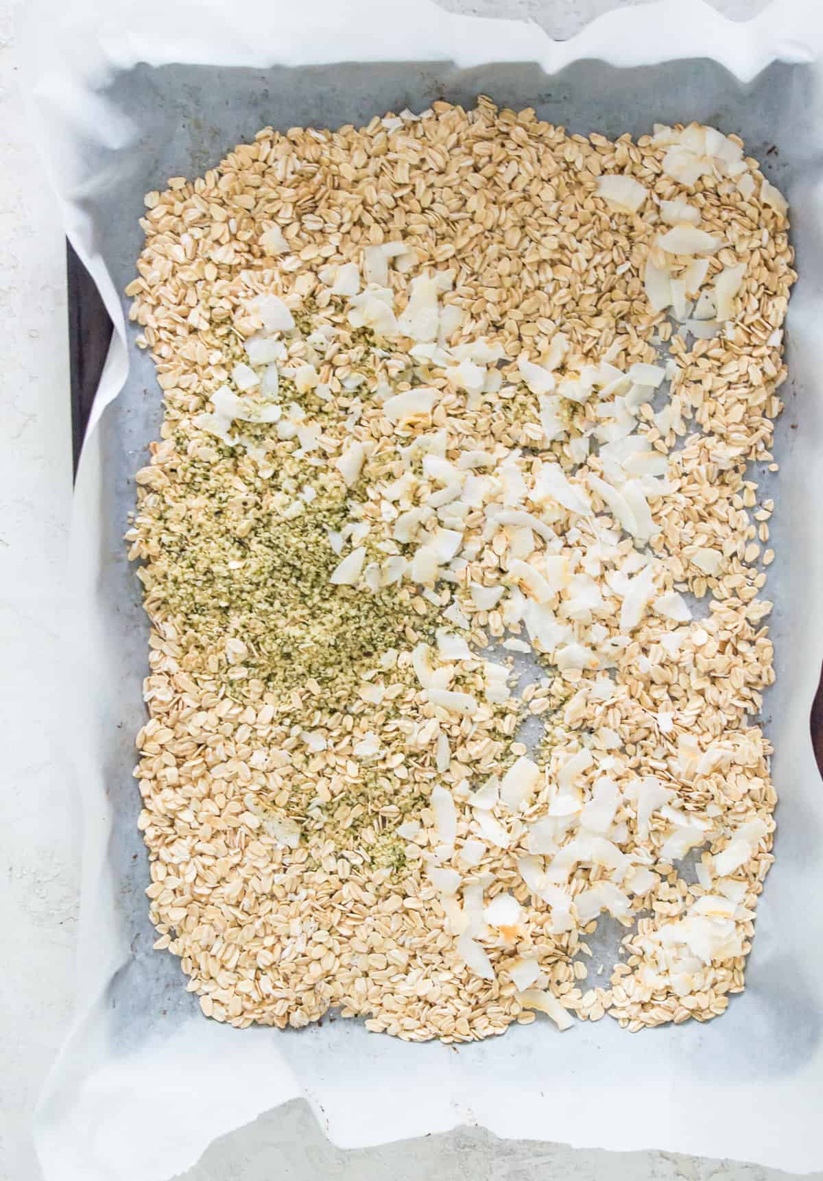 A baking sheet lined with parchment paper with oats and coconut flakes spread out over it. 