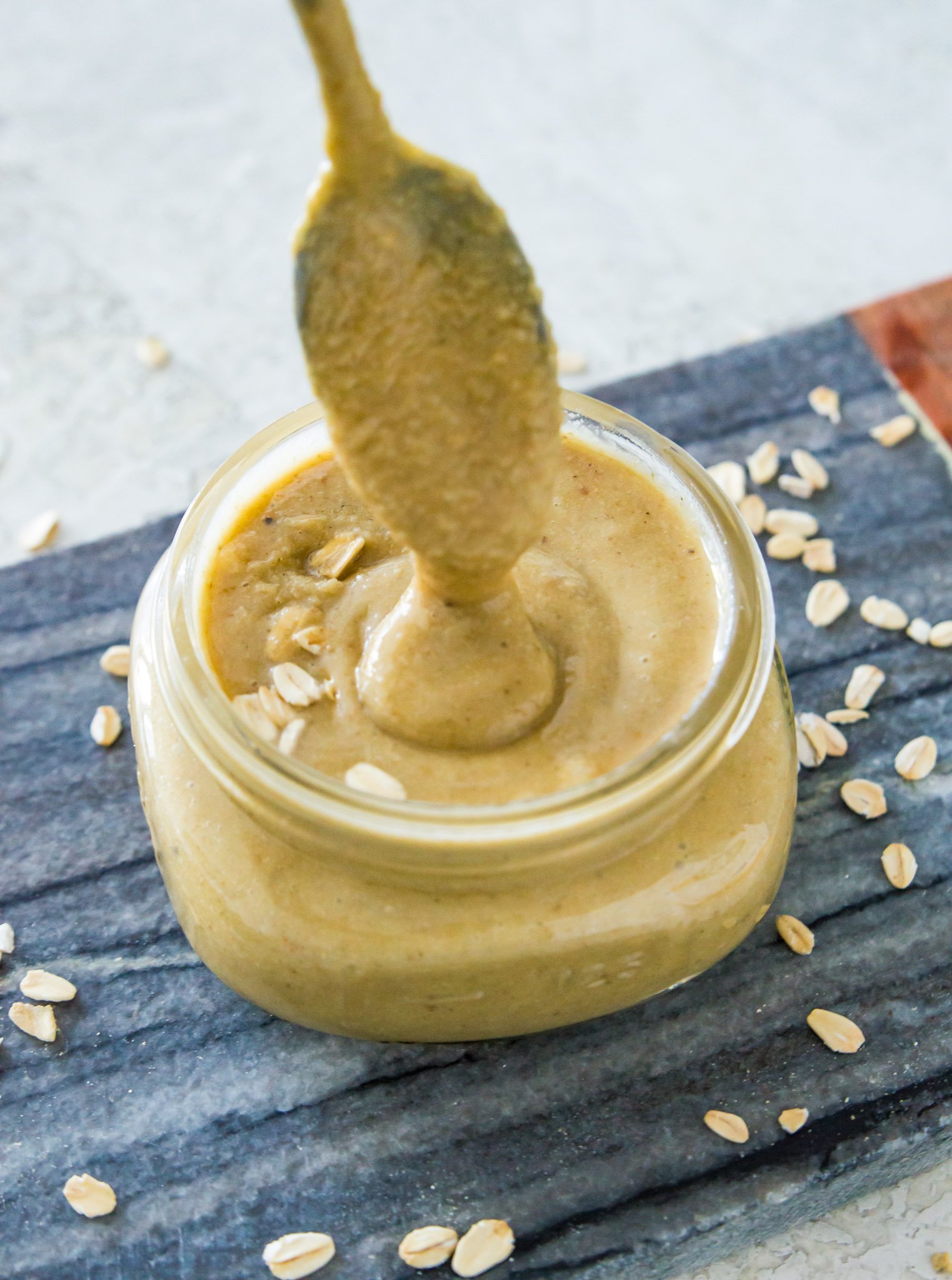 A jar of granola butter garnished with rolled oats with a spoon coated in the butter being pulled out of it.