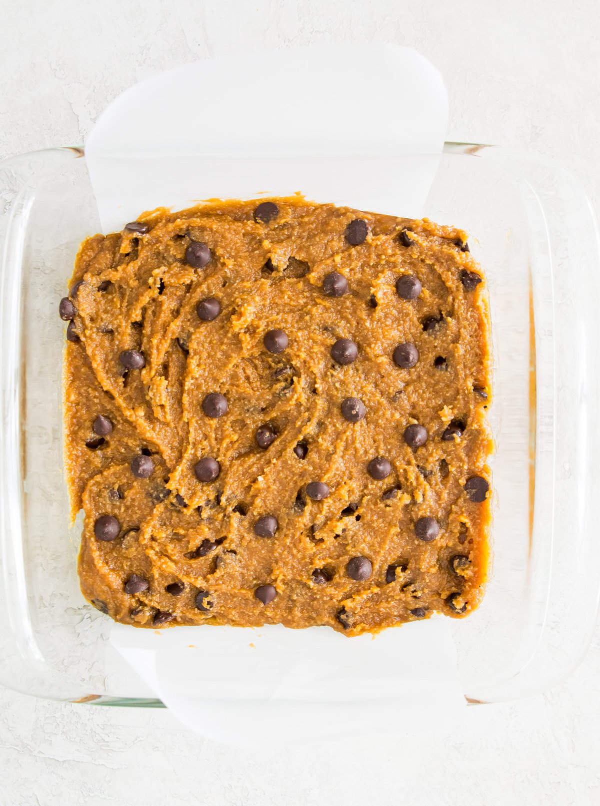Chocolate chip cookie dough spread out in a baking pan lined with parchment paper.